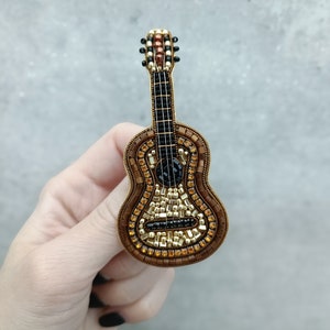 Könnte beinhalten: Eine Perlen-Gitarrenbrosche in Braun, Gold und Schwarz. Die Gitarre ist aus kleinen Perlen gefertigt und hat ein detailliertes Design.