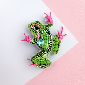 Può includere: Una spilla a forma di rana verde realizzata con perline, con accenti rosa e un occhio di cristallo trasparente. La spilla è su uno sfondo bianco e rosa.