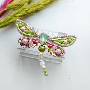 May include: A green and pink beaded dragonfly brooch with a crystal accent. The dragonfly has a long beaded tail.