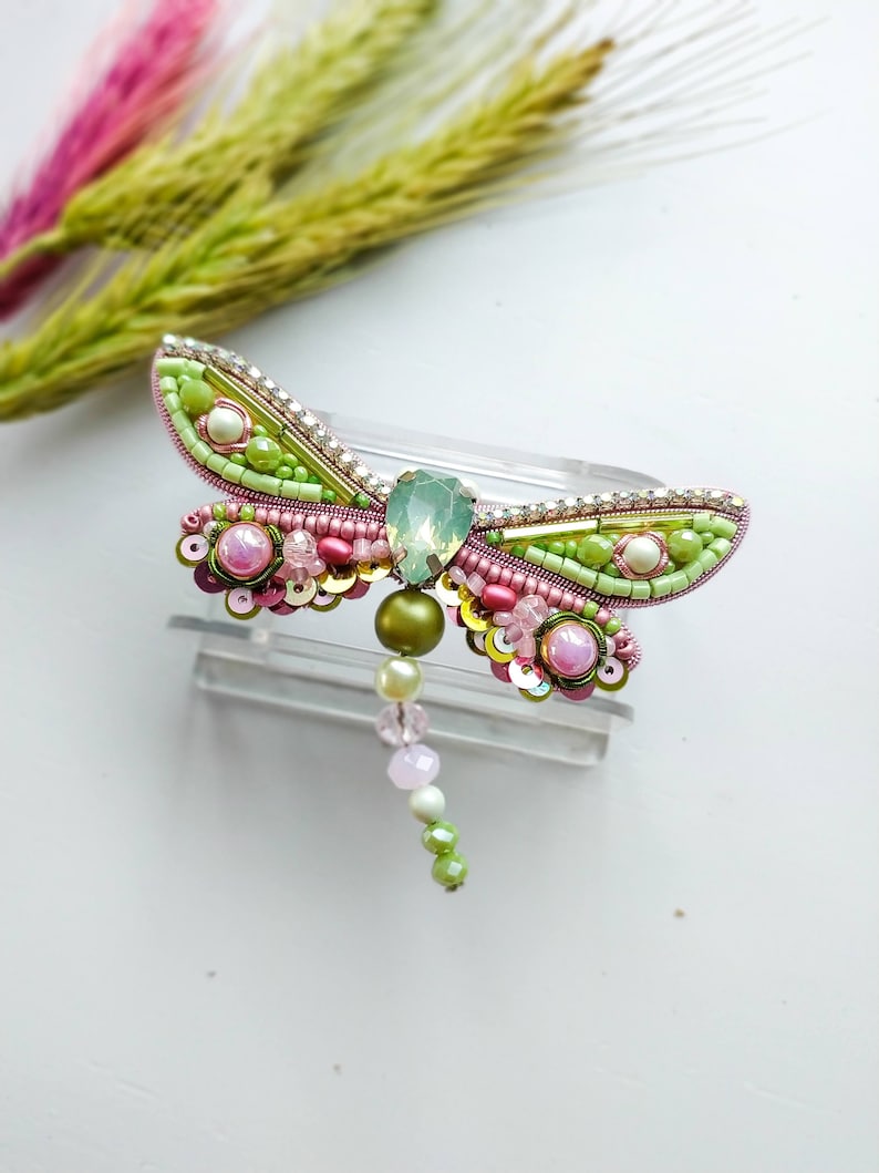 May include: A green and pink beaded dragonfly brooch with a crystal accent. The dragonfly has a long beaded tail.