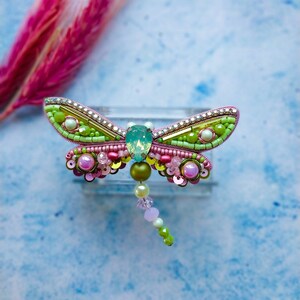 May include: A pink and green beaded dragonfly brooch with a light blue gem in the center. The dragonfly has a long beaded tail.