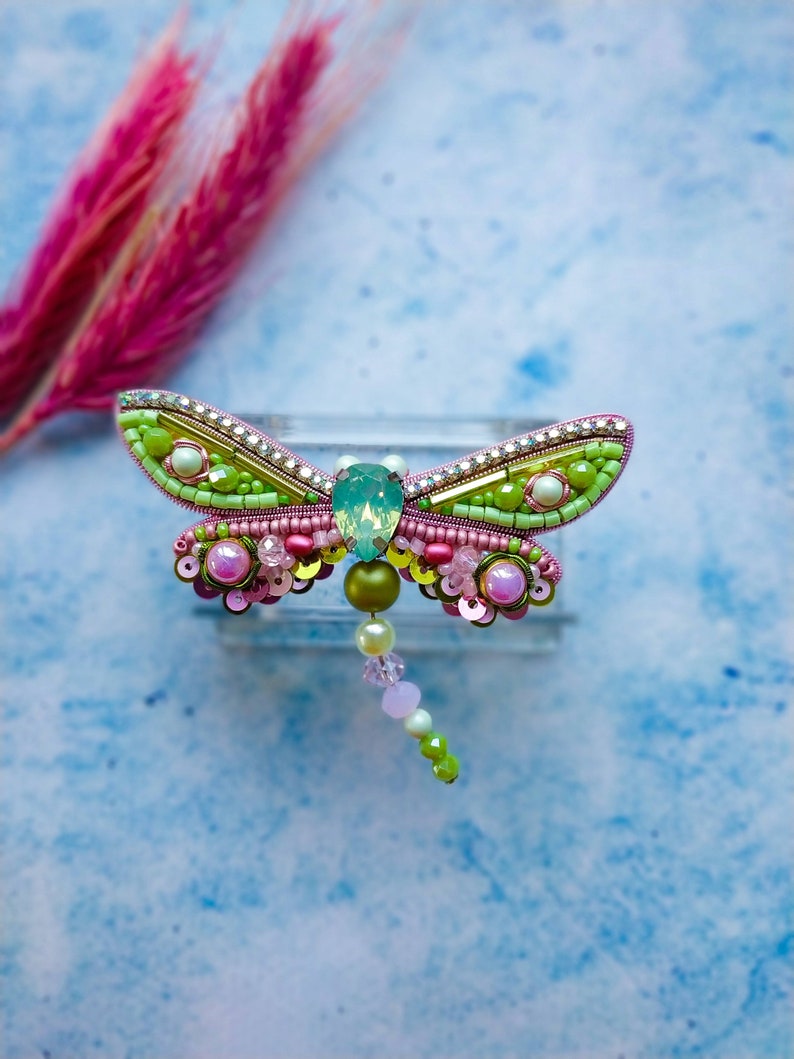 May include: A pink and green beaded dragonfly brooch with a light blue gem in the center. The dragonfly has a long beaded tail.