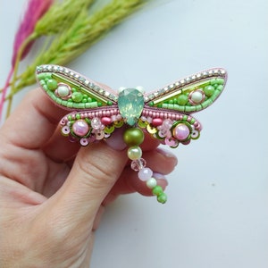 May include: A green and pink beaded dragonfly brooch with a clear crystal center. The dragonfly has a long beaded tail.