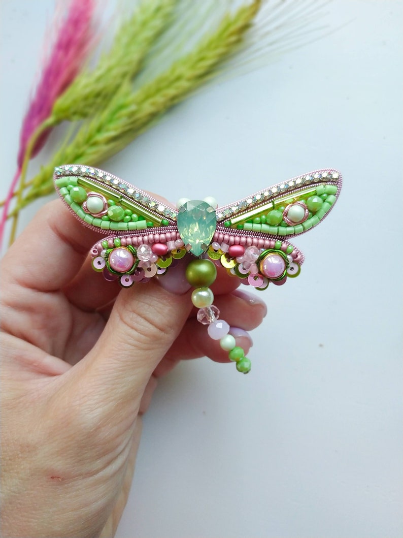 May include: A green and pink beaded dragonfly brooch with a clear crystal center. The dragonfly has a long beaded tail.