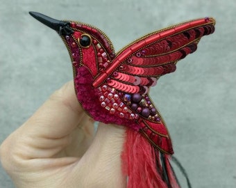 May include: A red hummingbird brooch with black feathers and sequins. The brooch is made with red velvet, beads, and sequins.