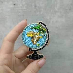 K&ouml;nnte beinhalten: Eine Perlen-Globus-Brosche mit einem blauen, gr&uuml;nen und gelben Design. Der Globus steht auf einem schwarzen Sockel mit einer goldenen Basis.