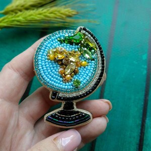 K&ouml;nnte beinhalten: Eine Perlen-Globus-Brosche mit blauer Basis, gelben und gr&uuml;nen Akzenten und einem schwarzen und goldenen Sockel.