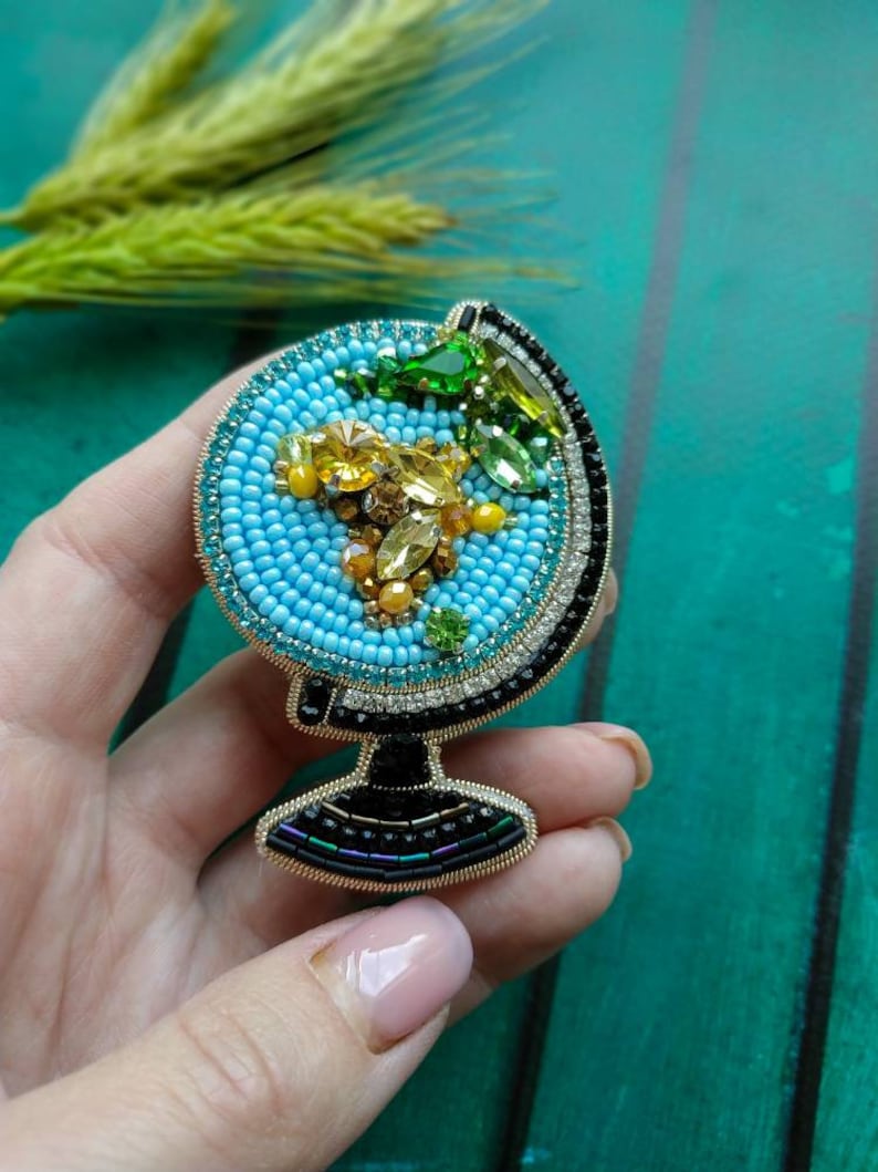 K&ouml;nnte beinhalten: Eine Perlen-Globus-Brosche mit blauer Basis, gelben und gr&uuml;nen Akzenten und einem schwarzen und goldenen Sockel.