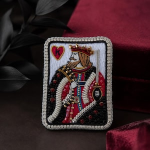 Puede incluir: Un broche rectangular con un diseño bordado de la carta del Rey de Corazones. La figura viste una túnica roja y negra con detalles dorados, una corona y sostiene una espada. El borde está adornado con cuentas plateadas y cristales claros.