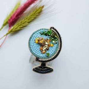 K&ouml;nnte beinhalten: Eine blaue und goldene Perlen-Globus-Brosche mit einem schwarzen Sockel. Der Globus ist mit bunten Perlen und Strasssteinen verziert.