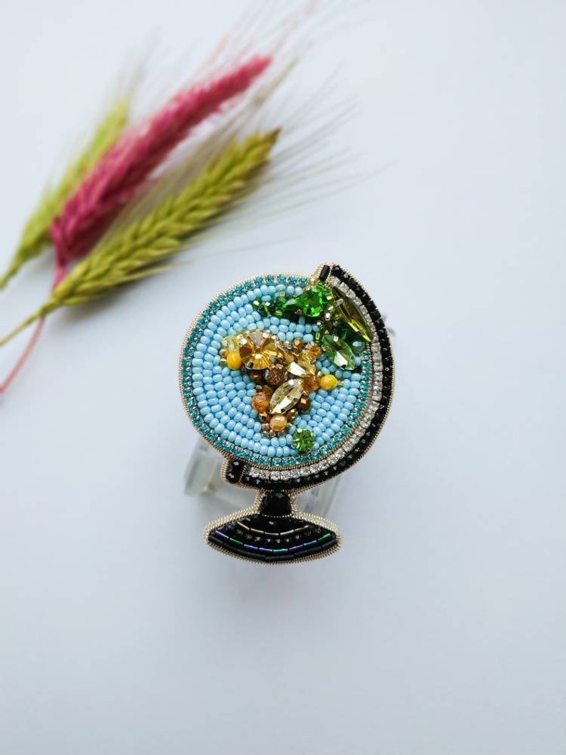 K&ouml;nnte beinhalten: Eine blaue und goldene Perlen-Globus-Brosche mit einem schwarzen Sockel. Der Globus ist mit bunten Perlen und Strasssteinen verziert.