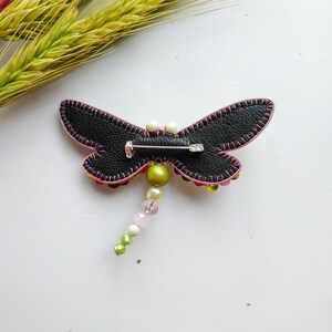 May include: A black leather dragonfly brooch with pink stitching and a beaded tail. The tail features green, white, and pink beads.