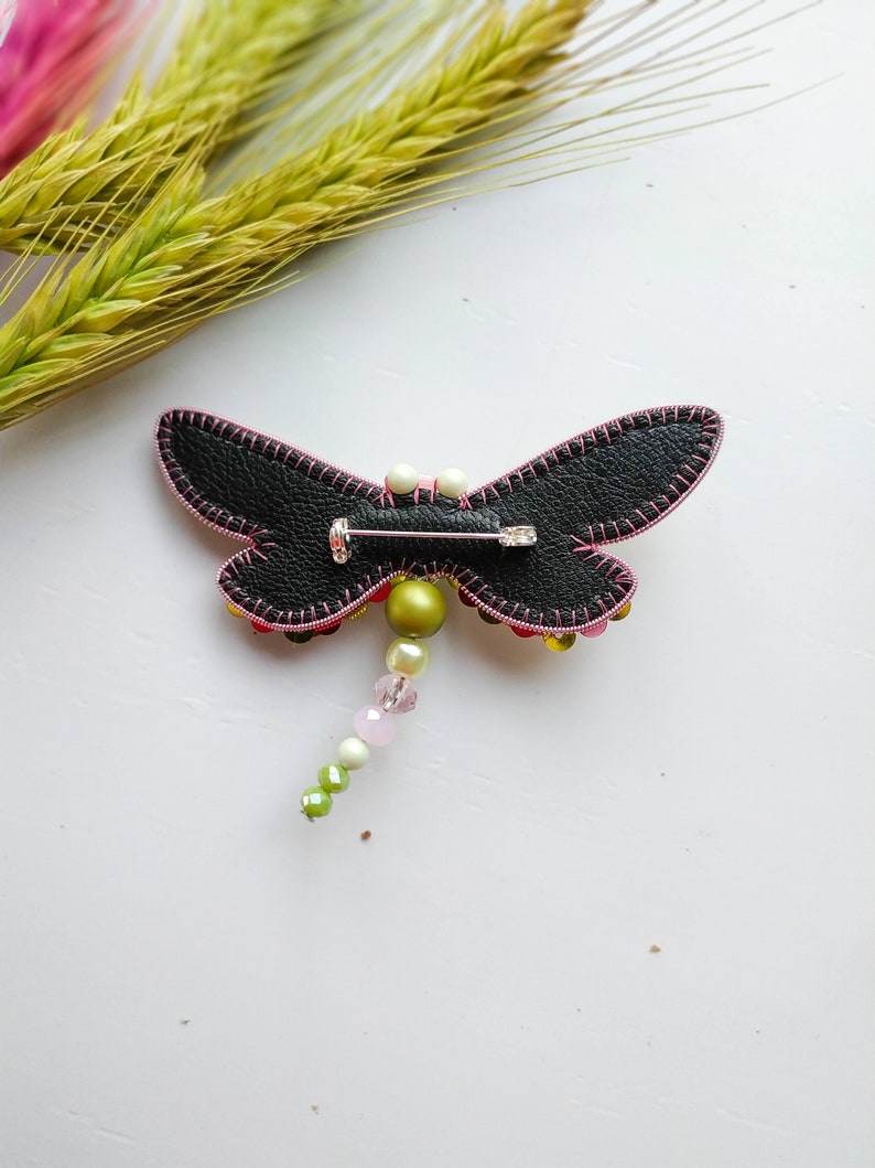 May include: A black leather dragonfly brooch with pink stitching and a beaded tail. The tail features green, white, and pink beads.