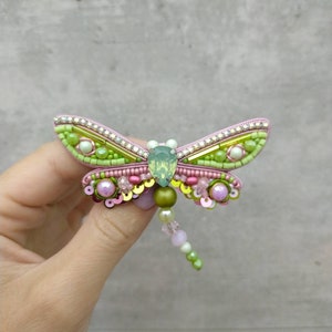 May include: A green and pink beaded dragonfly brooch with a clear crystal body. The wings are made of green and gold beads and sequins.