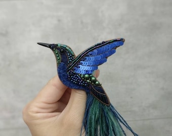 May include: A blue and green hummingbird brooch with iridescent sequins and feathers. The brooch is handmade and features a long tail of green feathers.