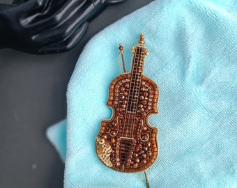 Op de afbeelding: Een bruine geperde viool broche met gouden accenten. De broche ligt op een lichtblauwe stoffen achtergrond.
