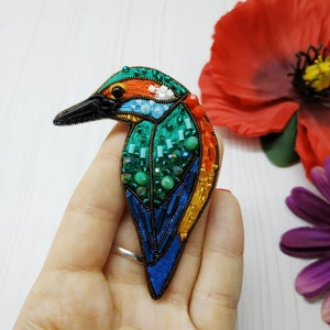 May include: A colorful beaded bird brooch with a green, blue, orange, and red design. The bird is facing left and has a black beak.