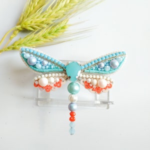 May include: A turquoise blue dragonfly brooch with white, pink, and orange beads. The dragonfly has a clear crystal body and a beaded tail.
