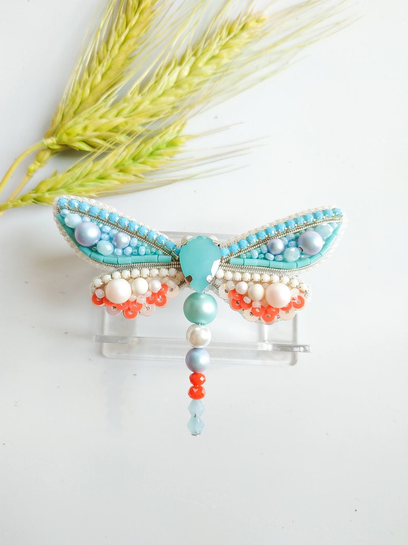 May include: A turquoise blue dragonfly brooch with white, pink, and orange beads. The dragonfly has a clear crystal body and a beaded tail.