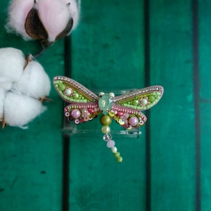 May include: A green and pink beaded dragonfly brooch with a clear crystal body and a beaded tail.
