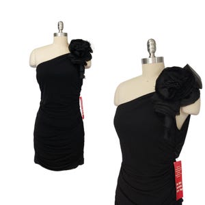 May include: A black one-shoulder dress with a ruched design. The dress features a large, decorative black flower on the shoulder. The dress is displayed on a mannequin.