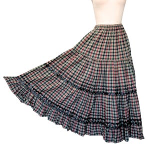 May include: A long, tiered skirt in a plaid pattern of red, green, and black on a cream background. The skirt has a ruffled hem with black trim and blue floral accents. The skirt is displayed on a mannequin.
