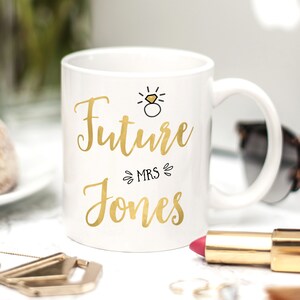 K&ouml;nnte beinhalten: Wei&szlig;e Keramiktasse mit goldenem Design, auf der "Future Mrs. Jones" steht, mit einer goldenen Ring- und Diamant-Illustration &uuml;ber dem Text.