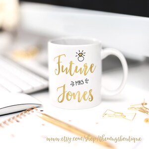 K&ouml;nnte beinhalten: Wei&szlig;e Keramiktasse mit goldenem Design. Die Tasse tr&auml;gt die Aufschrift "Future Mrs. Jones" mit einem goldenen Ring und einer Diamantgrafik &uuml;ber dem Text.