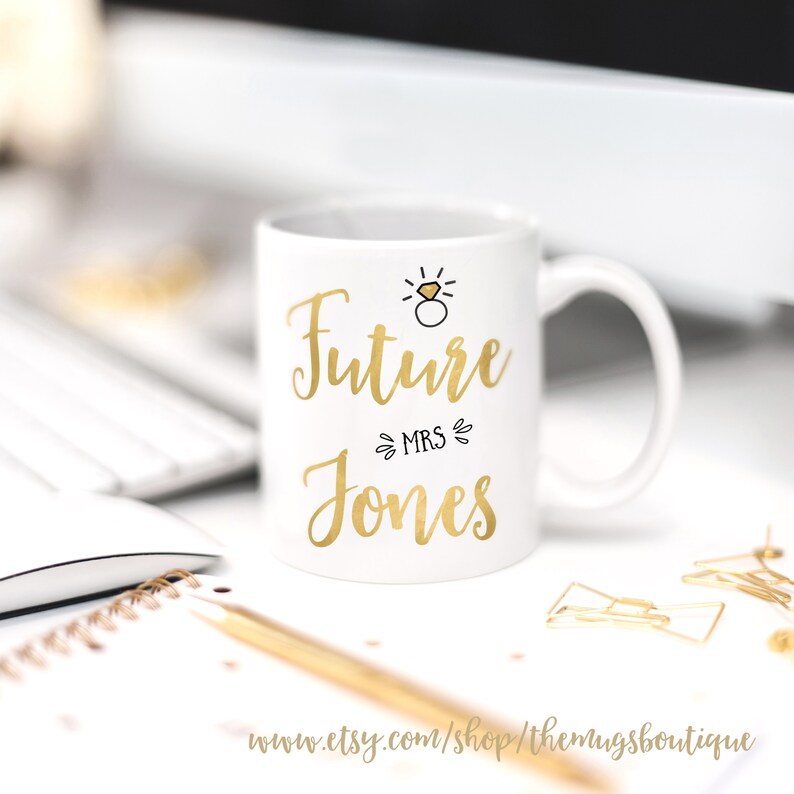 K&ouml;nnte beinhalten: Wei&szlig;e Keramiktasse mit goldenem Design. Die Tasse tr&auml;gt die Aufschrift "Future Mrs. Jones" mit einem goldenen Ring und einer Diamantgrafik &uuml;ber dem Text.