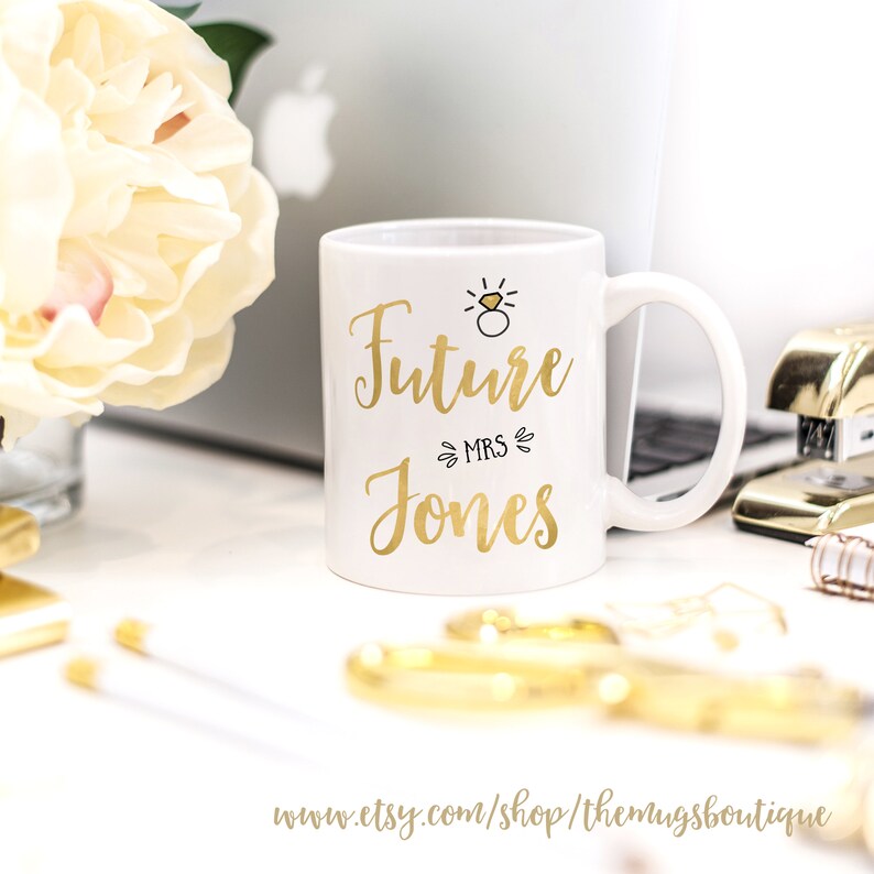 K&ouml;nnte beinhalten: Wei&szlig;e Keramiktasse mit goldenem Design, auf der "Future Mrs. Jones" steht, mit einem kleinen Ring und einer Diamantgrafik &uuml;ber dem Text.