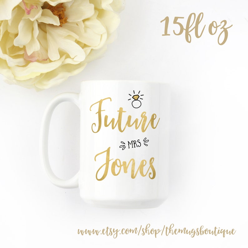 K&ouml;nnte beinhalten: Wei&szlig;e Keramiktasse mit goldener Aufschrift "Future Mrs. Jones" mit einer kleinen Ringzeichnung &uuml;ber dem Text. Die Tasse ist 15 fl oz.