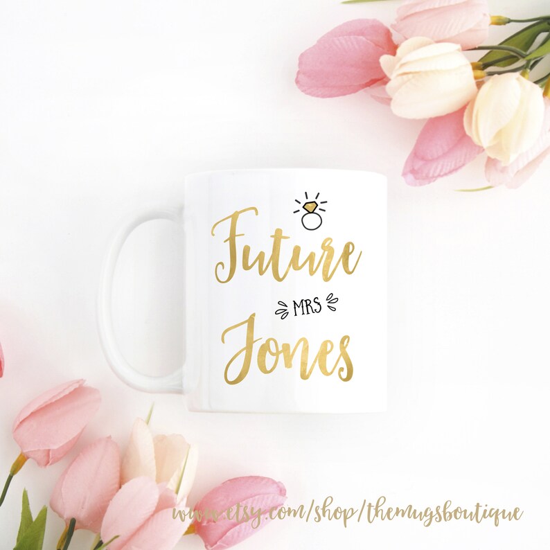 K&ouml;nnte beinhalten: Wei&szlig;e Keramiktasse mit goldenem Design, auf der "Future Mrs. Jones" steht, mit einer kleinen Ring- und Diamant-Illustration &uuml;ber dem Text.