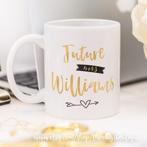Puede incluir: Taza de caf&eacute; de cer&aacute;mica blanca con una inscripci&oacute;n dorada que dice "Future Mrs. Williams" con una pancarta negra que dice "mrs" y una flecha dorada con un coraz&oacute;n.