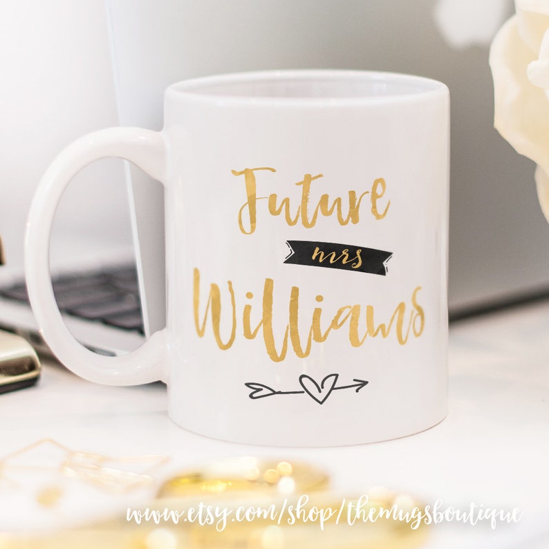 Puede incluir: Taza de caf&eacute; de cer&aacute;mica blanca con una inscripci&oacute;n dorada que dice "Future Mrs. Williams" con una pancarta negra que dice "mrs" y una flecha dorada con un coraz&oacute;n.