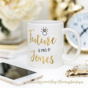 K&ouml;nnte beinhalten: Wei&szlig;e Keramiktasse mit goldener Aufschrift "Future Mrs. Jones" und einer goldenen Illustration eines Rings mit Diamant &uuml;ber dem Text.