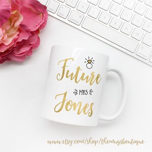 K&ouml;nnte beinhalten: Wei&szlig;e Keramiktasse mit goldener Aufschrift "Future Mrs. Jones" mit einer kleinen goldenen Ring- und Diamant-Illustration &uuml;ber dem Text.