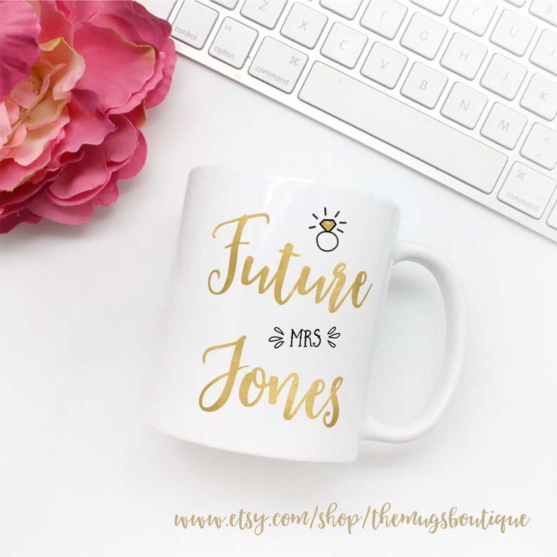 K&ouml;nnte beinhalten: Wei&szlig;e Keramiktasse mit goldener Aufschrift "Future Mrs. Jones" mit einer kleinen goldenen Ring- und Diamant-Illustration &uuml;ber dem Text.