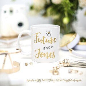 K&ouml;nnte beinhalten: Wei&szlig;e Keramiktasse mit goldenem Design, auf der "Future Mrs. Jones" steht, mit einer goldenen Ring- und Diamant-Illustration &uuml;ber dem Text.