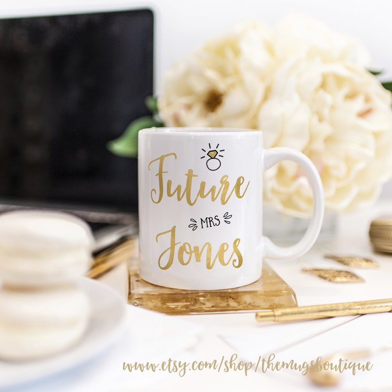 K&ouml;nnte beinhalten: Wei&szlig;e Keramiktasse mit goldenem Design, auf der "Future Mrs. Jones" steht, mit einer goldenen Ring- und Diamant-Illustration &uuml;ber dem Text.