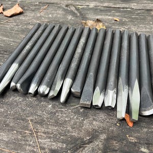 Puede incluir: Una colecci&oacute;n de quince herramientas manuales de metal gris oscuro, que incluyen varios punzones y cinceles con puntas de plata pulida. Las herramientas est&aacute;n dispuestas en una fila paralela, mostrando diferentes formas de punta como planas, puntiagudas y c&oacute;nicas.