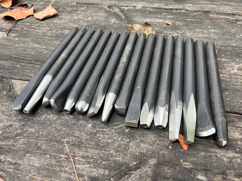 Puede incluir: Una colecci&oacute;n de quince herramientas manuales de metal gris oscuro, que incluyen varios punzones y cinceles con puntas de plata pulida. Las herramientas est&aacute;n dispuestas en una fila paralela, mostrando diferentes formas de punta como planas, puntiagudas y c&oacute;nicas.