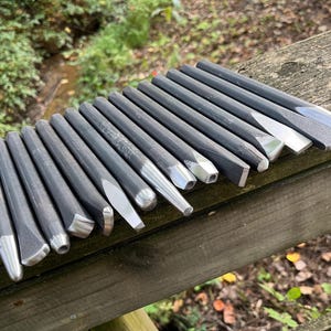 Puede incluir: Una colecci&oacute;n de quince herramientas manuales de metal gris oscuro, que incluyen varios cinceles y punzones con puntas plateadas pulidas. Las herramientas est&aacute;n dispuestas en una fila paralela, mostrando diferentes formas de cabeza como planas, puntiagudas y redondeadas.