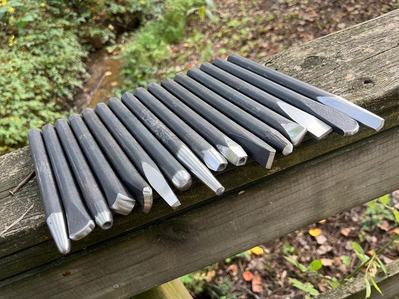 Puede incluir: Una colecci&oacute;n de quince herramientas manuales de metal gris oscuro, que incluyen varios cinceles y punzones con puntas plateadas pulidas. Las herramientas est&aacute;n dispuestas en una fila paralela, mostrando diferentes formas de cabeza como planas, puntiagudas y redondeadas.