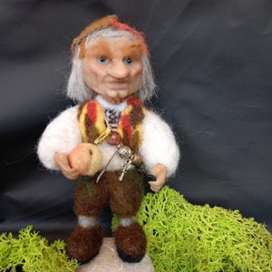 Op de afbeelding: Een fantasievolle vilten gnoompup met een witte baard, die een rood-bruine hoed, een wit shirt, een bruine broek draagt en een klein perzikkleurig fruit vasthoudt. De gnoom staat op een rots met groen mos.