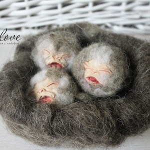 May include: Three small felt sculptures of baby faces with closed eyes, nestled together in a bed of gray wool. The sculptures have realistic features and are made with a soft, fuzzy texture.