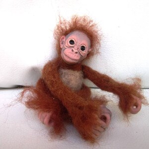 Puede incluir: Una pequeña muñeca de orangután hecha a mano con rasgos realistas, incluyendo pelaje marrón, rostro rosado y ojos grandes y oscuros. La muñeca está sentada sobre una superficie blanca.
