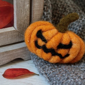 May include: A felt orange pumpkin with a black jack-o'-lantern face, sitting on a grey knitted fabric.