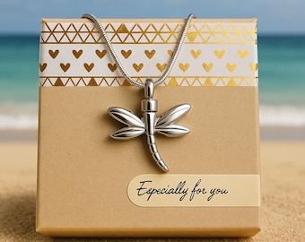 May include: A silver dragonfly pendant necklace displayed on a gift box with gold heart and triangle patterns. The box has a label that reads "Especially for you". The background shows a beach and ocean.