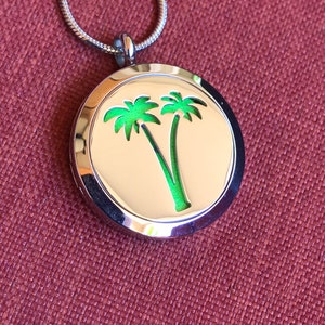 May include: A silver-colored pendant necklace with a circular charm featuring a green palm tree design against a cream-colored background. The necklace has a thin, snake chain and is displayed on a maroon fabric surface.