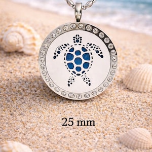 May include: Silver pendant necklace with a round charm featuring a blue and white sea turtle design. The charm is 2.5 cm in diameter and is surrounded by small, clear crystals. The necklace is displayed on a sandy beach.